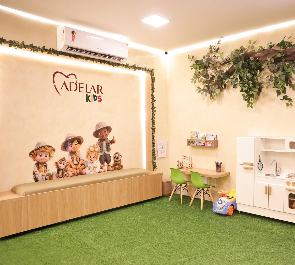Sala de espera da Adelar Kids: painel com o logo e personagens exploradores, banco acolchoado, mesinha com cadeiras infantis, cozinha de brinquedo, galho com folhagem no teto e piso de grama sintética